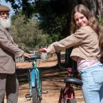 Fotografia de jovem sorridente e homem idoso de barba branca, cada um com uma bicicleta em caminho arborizado.
