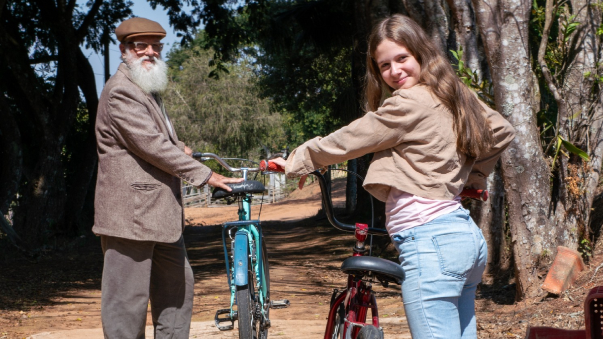 Fotografia de jovem sorridente e homem idoso de barba branca, cada um com uma bicicleta em caminho arborizado.