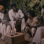 Fotografia de pessoas negras em roupas brancas de época, reunidas ao redor de uma mesa à luz de velas.