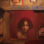 Cena teatral alaranjada com o rosto de uma jovem em uma tela e dois retratos abaixo.