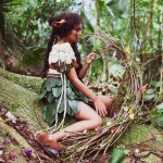 Fotografia de menina em traje de folhas e flores sentada na floresta, segurando uma guirlanda de galhos.