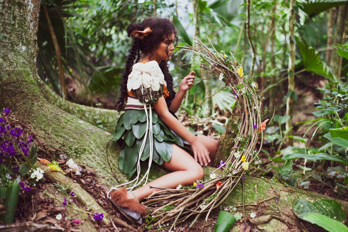 Fotografia de menina em traje de folhas e flores sentada na floresta, segurando uma guirlanda de galhos.
