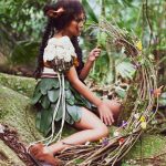 Fotografia de uma menina sentada em um grande ninho de galhos no meio da floresta.