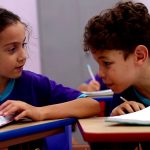 Fotografia de duas crianças uniformizadas conversando sobre cadernos em uma sala de aula.