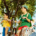 Fotografia de três meninos com bicicletas sob uma árvore, atentos a algo fora da cena.