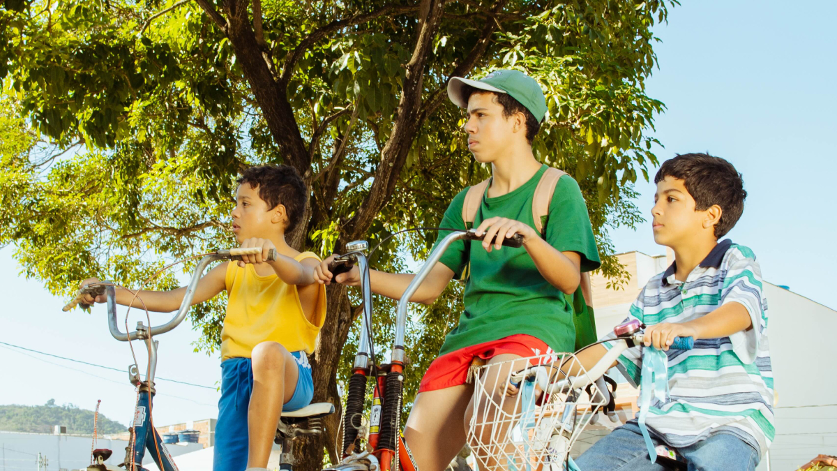 Fotografia de três meninos com bicicletas sob uma árvore, atentos a algo fora da cena.