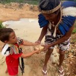 Fotografia de homem e menino indígenas, com adornos de penas, segurando juntos uma pedra em terreno lamacento.