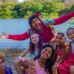 Fotografia de crianças e adulto sorridentes na margem de um rio, cercados por vegetação.