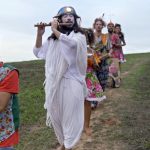 Fotografia de crianças e adultos com roupas brancas caminhando por um campo, com uma criança de vermelho à frente.