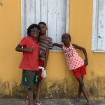 Fotografia de três crianças negras apoiadas em uma parede amarela, usando roupas coloridas.