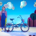 Animação de uma bicicleta ao centro, ao seu lado uma menina de trança, nuvens e formas urbanas coloridas.