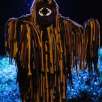 Fotografia de uma figura cênica coberta por longas tiras marrons, iluminada por luz azul.