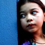Fotografia em close de uma menina olhando de lado diante de uma parede azul.