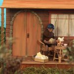 Animação em stop motion mostra personagem diante de uma pequena casa de madeira, cercada por vegetação.