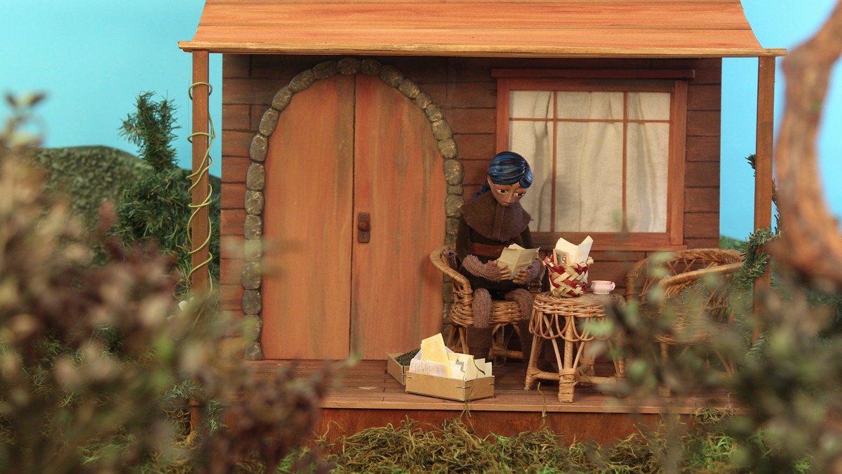 Animação em stop motion mostra personagem diante de uma pequena casa de madeira, cercada por vegetação.