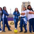 Fotografia mostra cinco meninas posando sorridentes em um campo de futebol, com uma bola amarela aos pés.