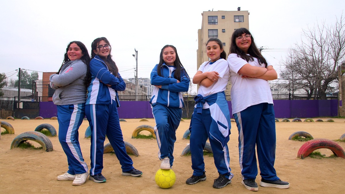Fotografia mostra cinco meninas posando sorridentes em um campo de futebol, com uma bola amarela aos pés.