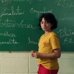 Fotografia de uma criança de camiseta amarela diante de uma lousa coberta por palavras escritas a giz.