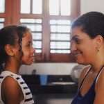 Fotografia de mãe e filha sorrindo frente a frente em uma sala iluminada.