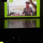 Fotografia de auditório escuro com tela exibindo homem ao ar livre e intérprete de Libras, com reflexo verde no palco.