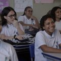 Fotografia mostra estudantes uniformizados sentados em uma sala de aula; uma jovem sorri em primeiro plano.