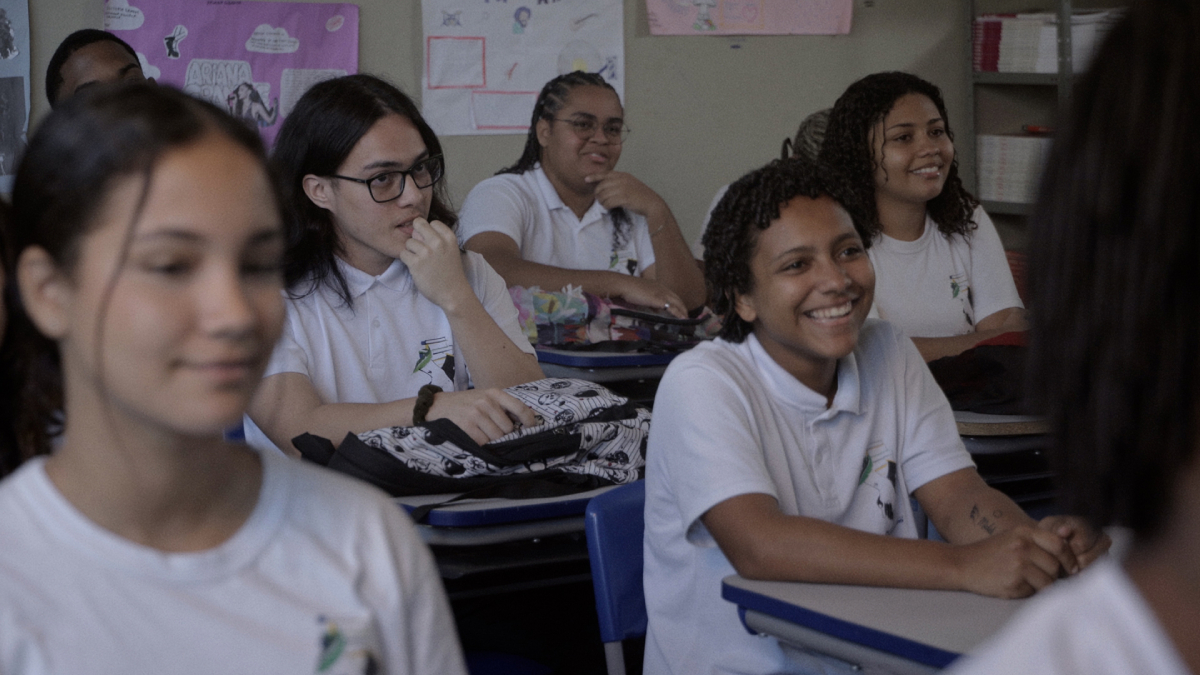 Fotografia mostra estudantes uniformizados sentados em uma sala de aula; uma jovem sorri em primeiro plano.