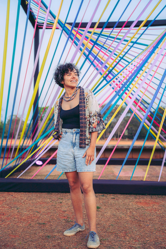 Fotografia de uma mulher sorridente, de cabelos curtos e cacheados, em pé diante de uma instalação artística formada por fios coloridos cruzados. Ela veste blusa preta, camisa estampada, shorts jeans e tênis.