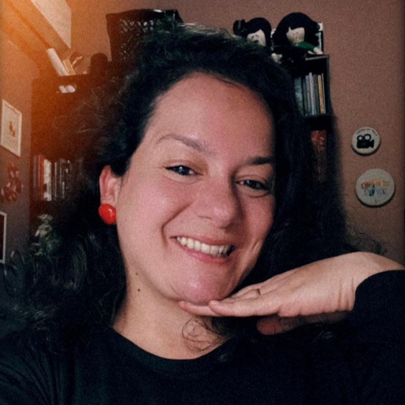 Fotografia de mulher sorridente, com cabelos escuros e brincos vermelhos, apoiando a mão sob o queixo.
