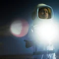 Fotografia de pessoa com traje de astronauta, iluminada por forte clarão no peito em ambiente escuro.