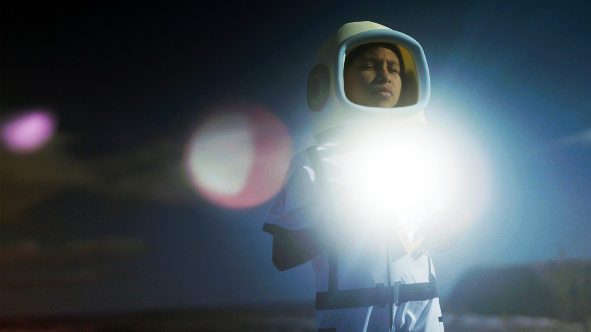 Fotografia de pessoa com traje de astronauta, iluminada por forte clarão no peito em ambiente escuro.