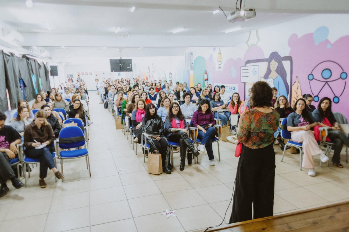 Fotografia de uma mulher falando de costas para dezenas de pessoas sentadas em uma sala com cadeiras azuis e paredes ilustradas.