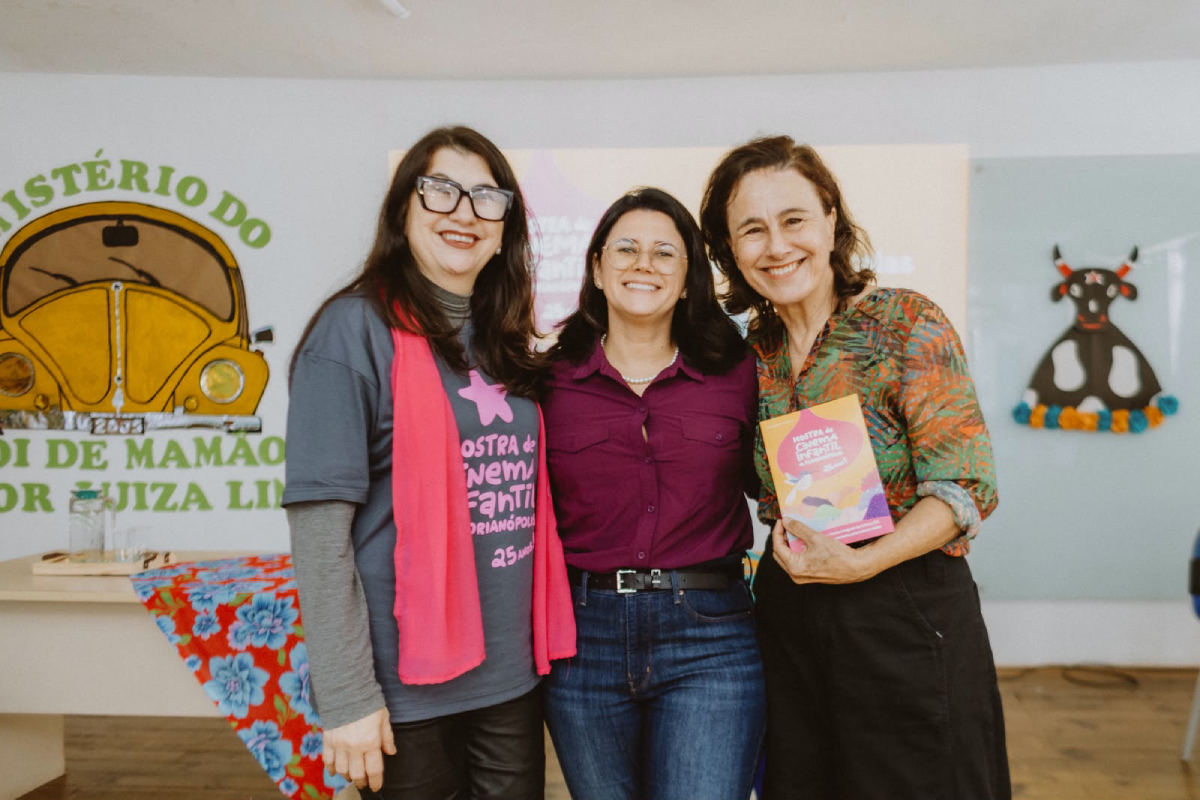 Fotografia de três mulheres sorridentes lado a lado; a da direita segura o programa da Mostra de Cinema Infantil.
