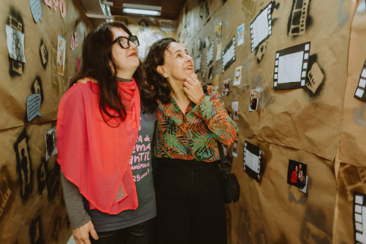 Fotografia de duas mulheres sorridentes observando fotografias e textos expostos nas paredes de um corredor.