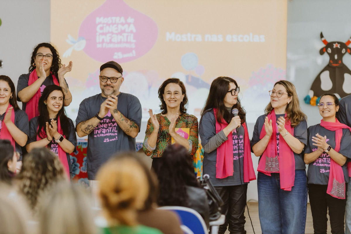 Fotografia de integrantes da Mostra de Cinema Infantil aplaudindo diante do público. Uma mulher segura o microfone; ao fundo, lê-se “Mostra nas escolas”.