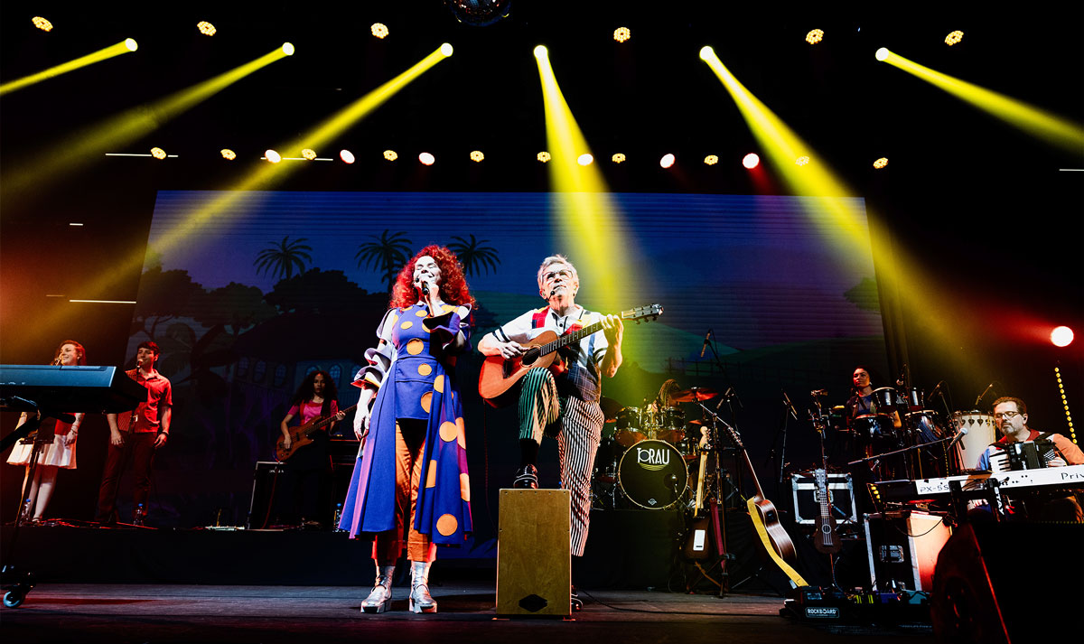 Fotografia de show da dupla Palavra Cantada em palco: uma mulher ruiva canta ao microfone ao lado de um homem tocando violão, enquanto músicos se apresentam ao fundo sob intensos feixes de luz amarela.