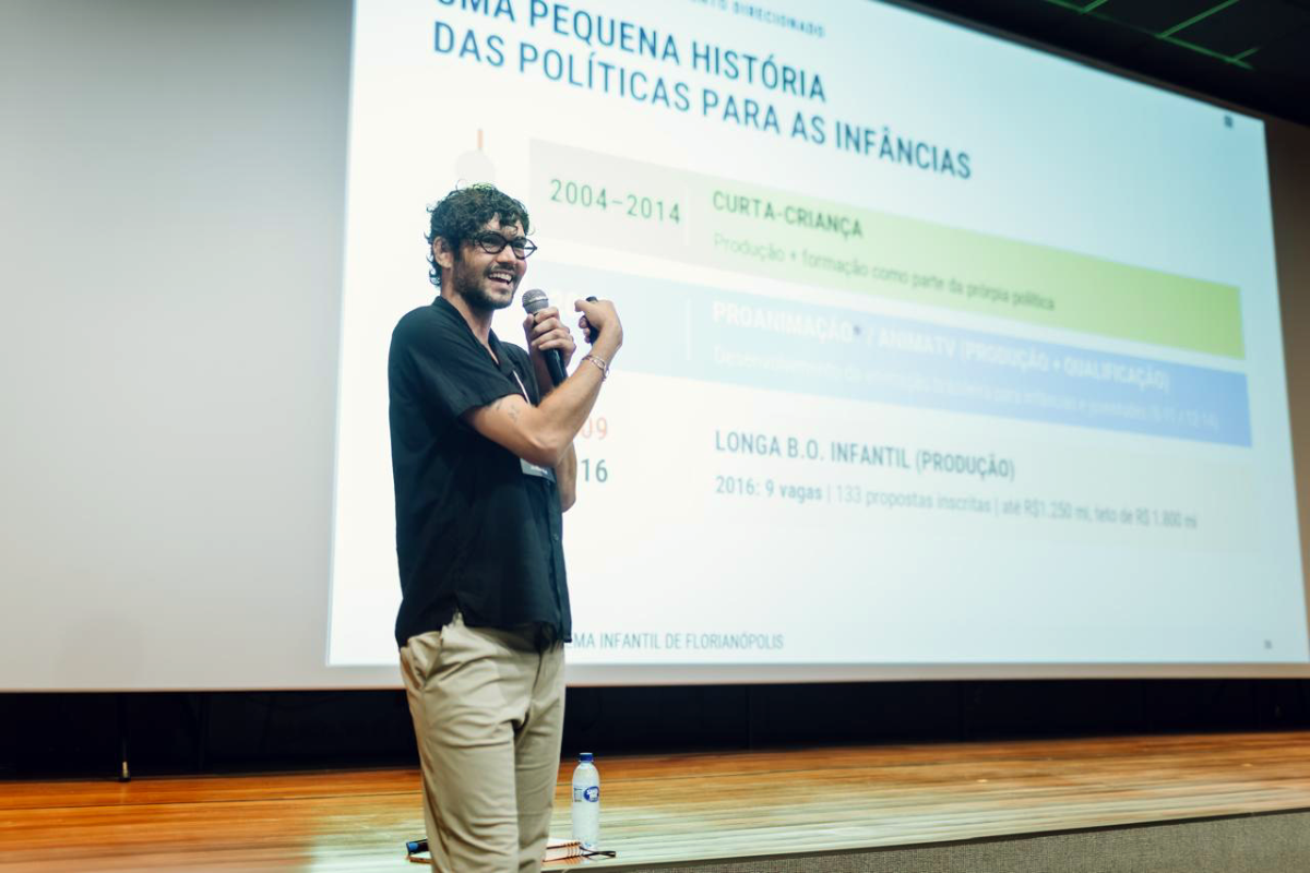 Fotografia de homem com óculos em um palco, sorrindo enquanto fala ao microfone diante de uma apresentação projetada com o título “Uma pequena história das políticas para as infâncias”